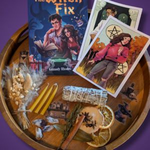 An autumn-themed Raincrest Coven's Mabon ritual box display featuring a signed paperback novel titled The Witch Fix, character art tarot cards, a sage smudge stick, a dried flower bouquet, crystals, mini yellow candles, stickers, and a simmer pot mixture on a round wooden tray.