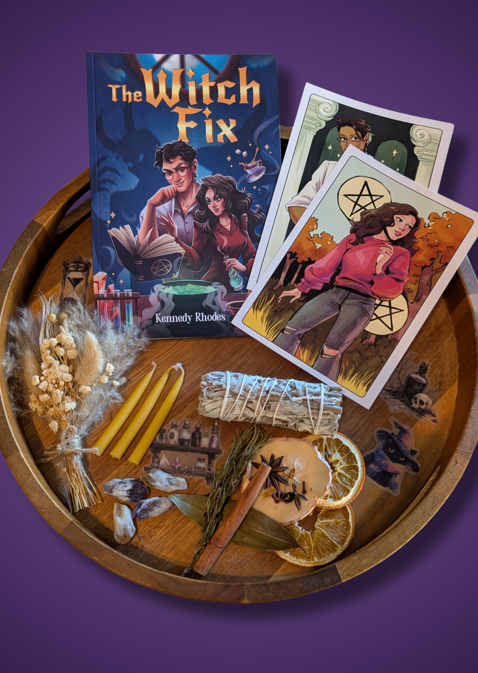 An autumn-themed Raincrest Coven's Mabon ritual box display featuring a signed paperback novel titled The Witch Fix, character art tarot cards, a sage smudge stick, a dried flower bouquet, crystals, mini yellow candles, stickers, and a simmer pot mixture on a round wooden tray.